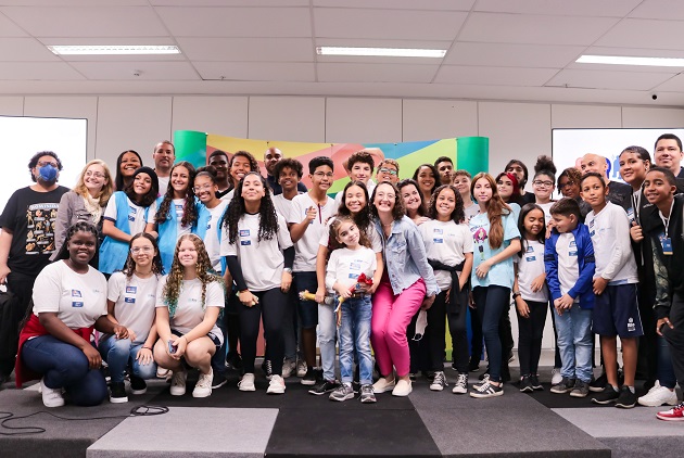 Fotografia colorida. Grupo de alunos e professores de escolas do Rio, posicionados em pé e à frente, agachados. Atrás, estão um painel colorido e dois telões em cada lateral .