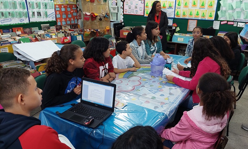 Fotografia. Em uma sala de aula, vários alunos estão sentados em volta de uma mesa conversando. Em primeiro plano, um aluno está de costas para a câmera e de frente para um computador. Ao fundo, uma professora em pé ouvindo os alunos.